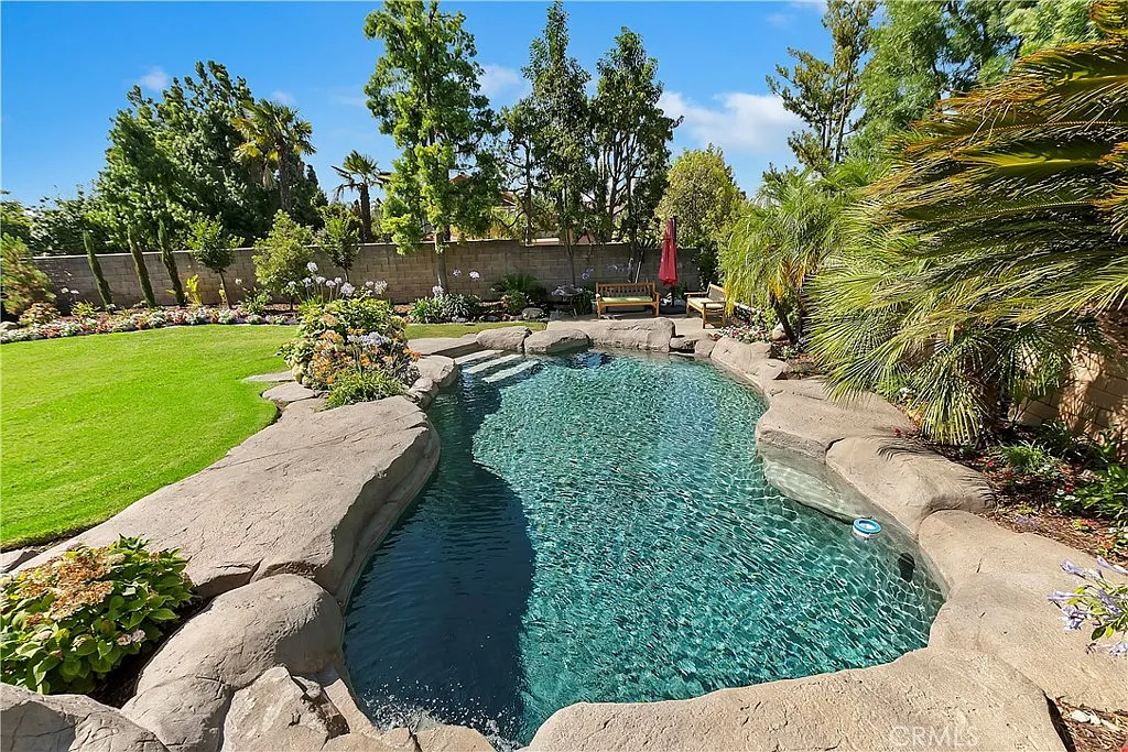 Landscaped backyard with curved pool, rocky edges, greenery, flowers, lounge chair, and umbrella under sunny sky