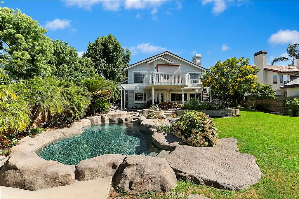 Backyard with swimming pool, rocks, plants, trees, and two-story house in background