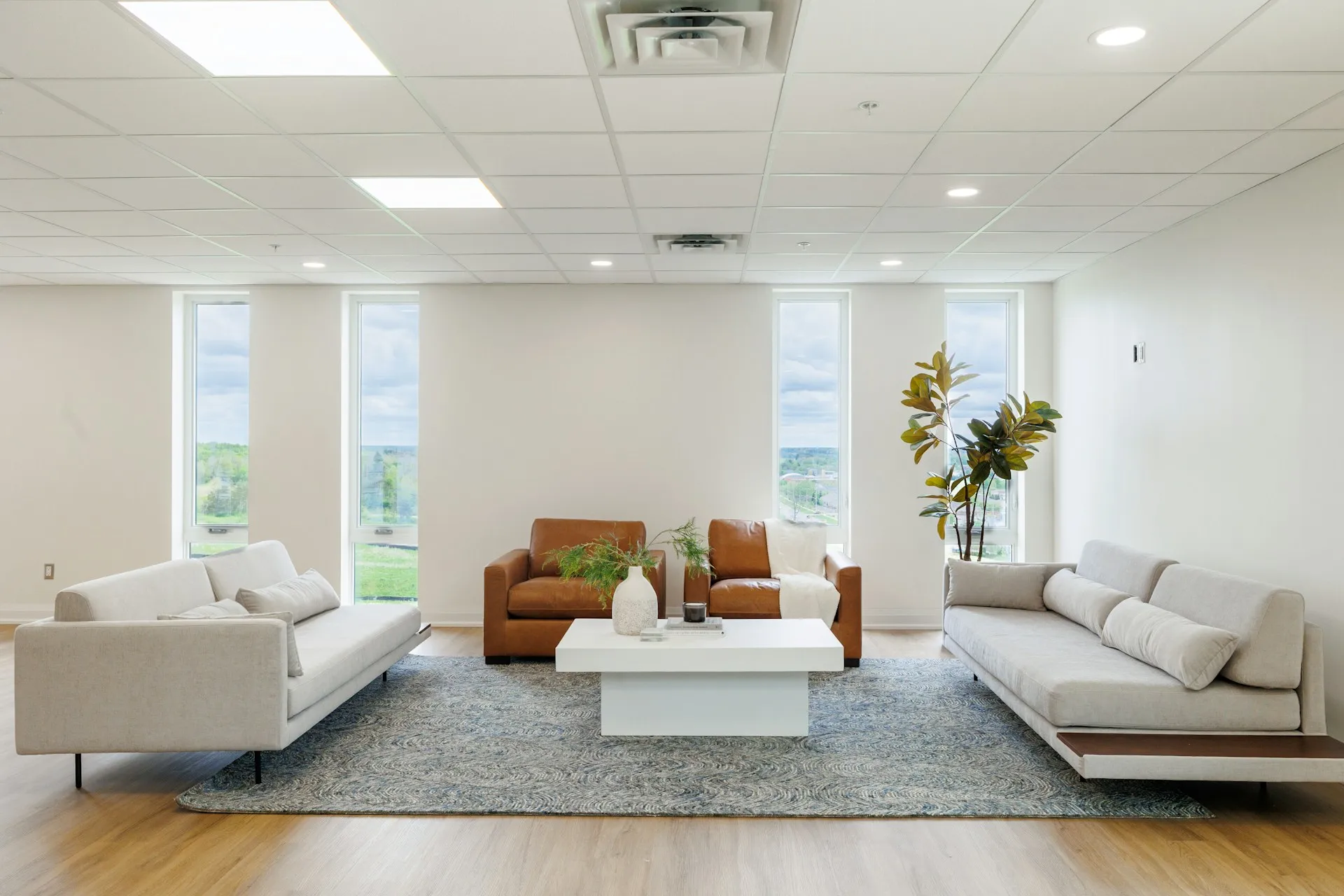 A living room with two sofas, an armchair, coffee table, plant, and large windows with light wood flooring.