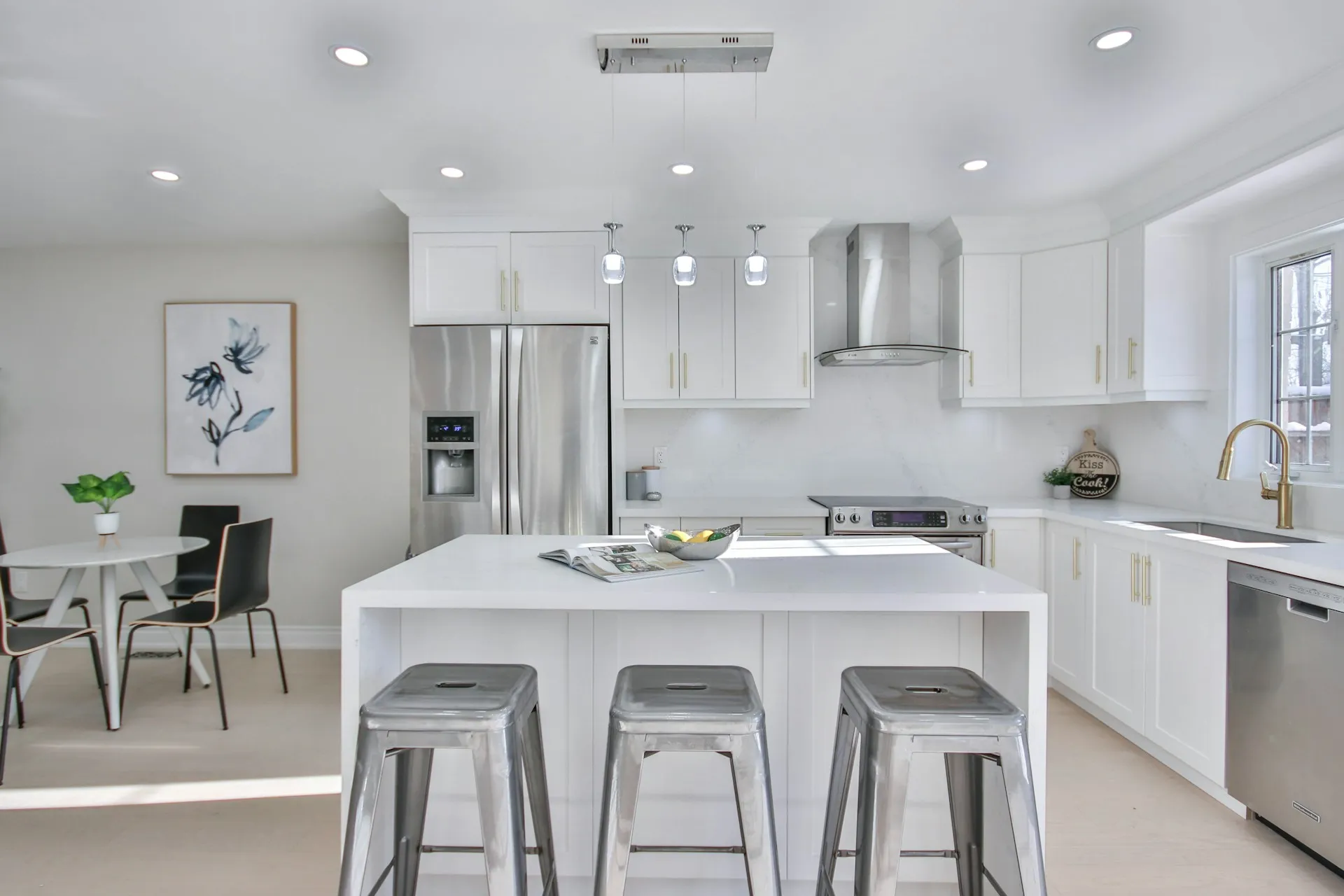 A kitchen with white cabinets, stainless steel appliances, island with bar stools, and a small dining table with plants.