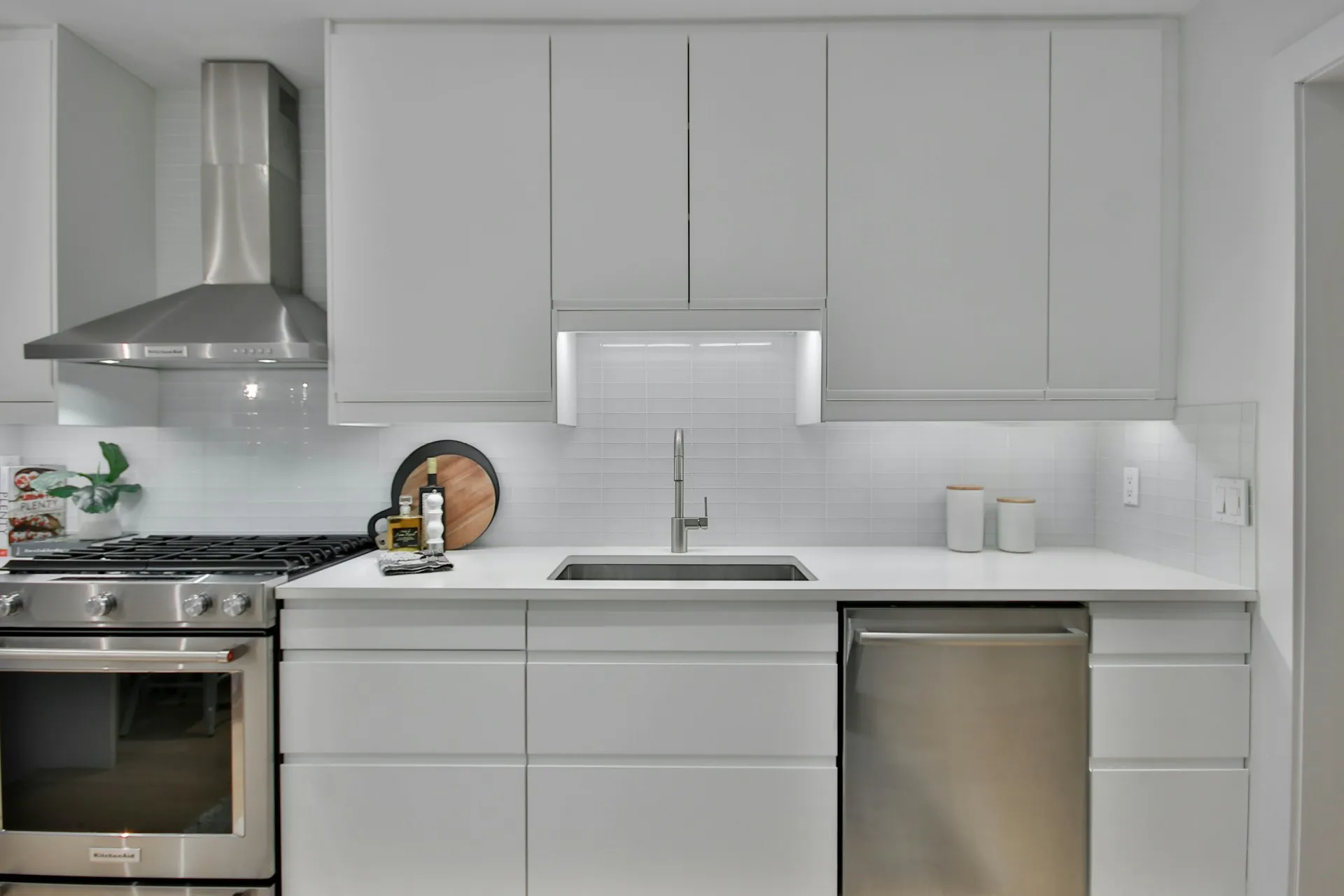 A kitchen with gray cabinets, stainless steel fridge, gas stove, sink, and white countertops.