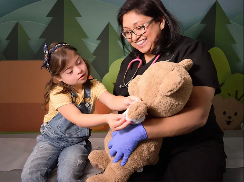 A child in overalls bandaging a stuffed animal with a caregiver wearing gloves and a stethoscope assisting.