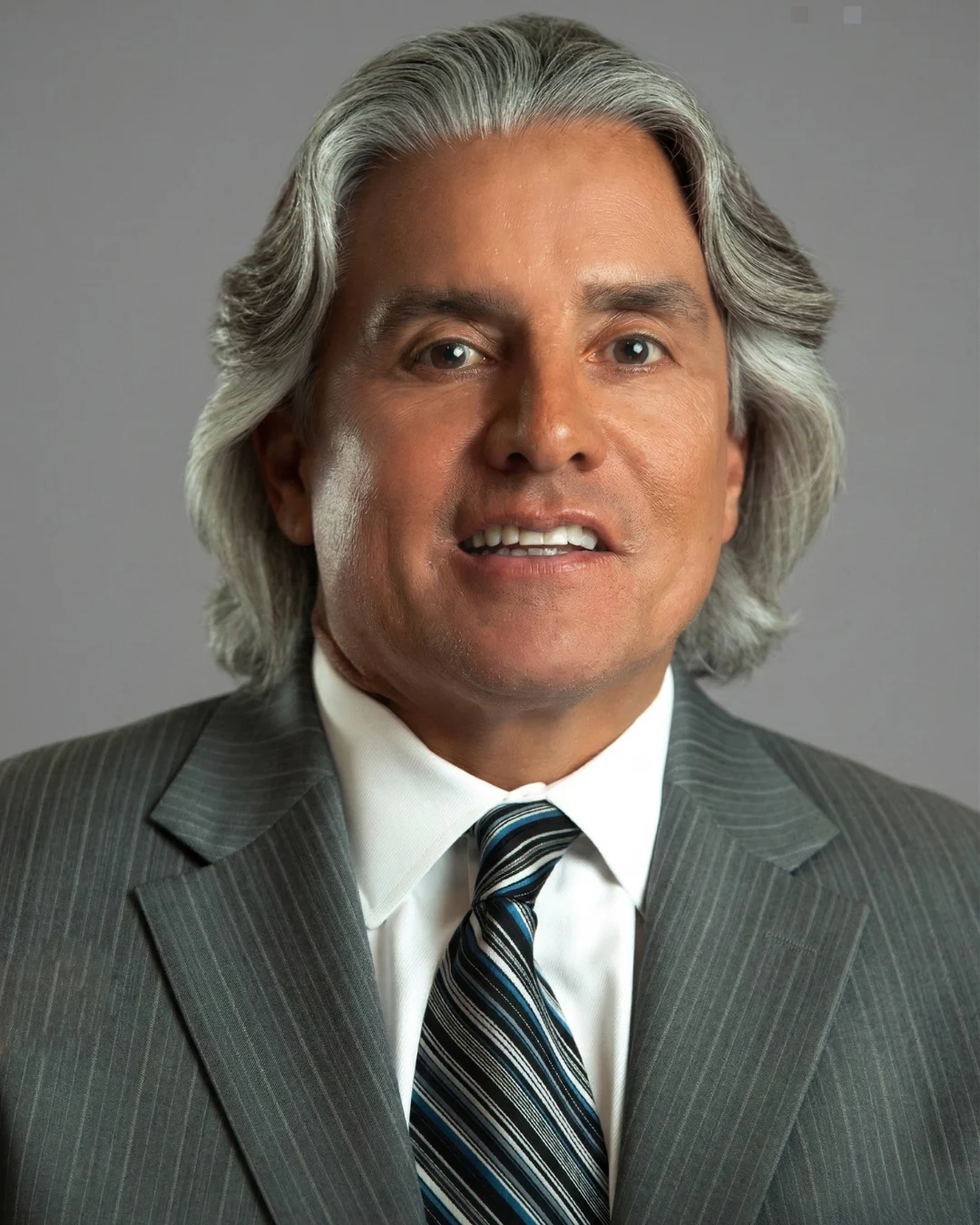 Smiling man with silver hair wearing gray suit, white shirt, and striped tie on plain gray background