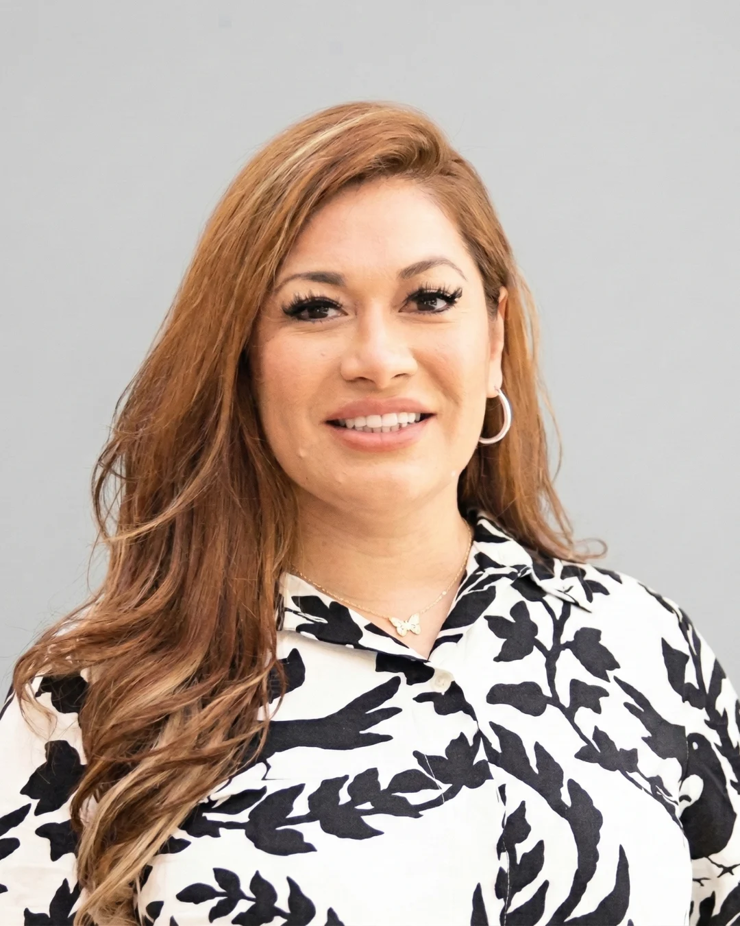 Woman with long wavy brown hair smiling at camera, wearing black and white patterned shirt and hoop earrings, plain gray background