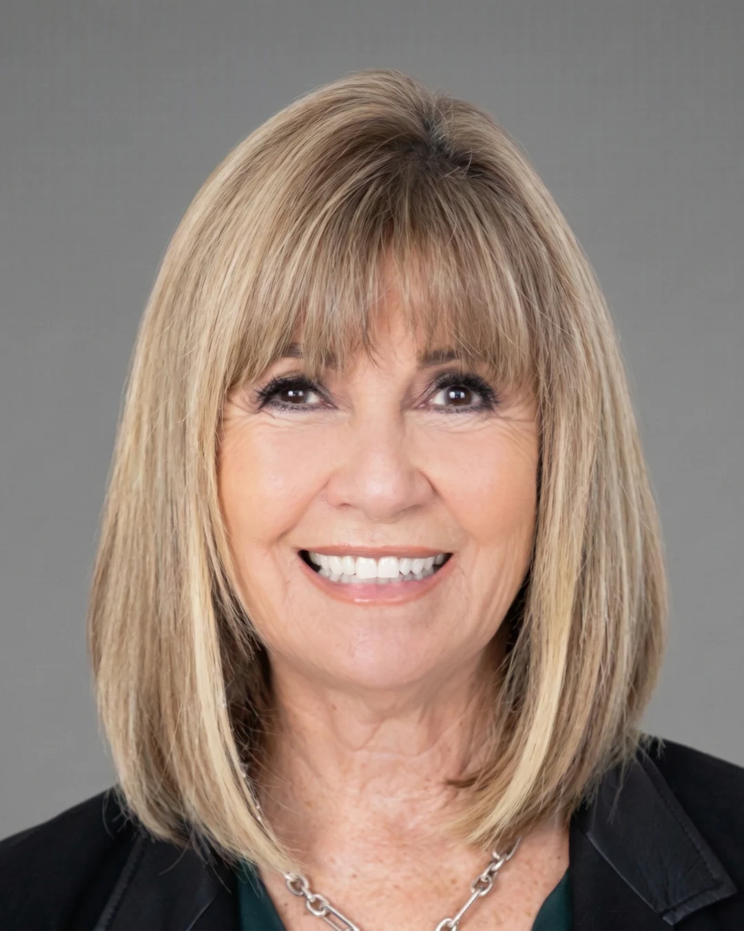 Woman with shoulder-length blonde hair and bangs smiling, wearing dark blazer and necklace on plain gray background