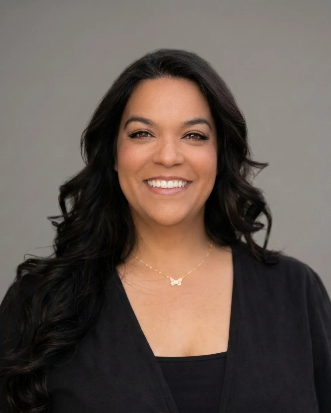 Woman with long dark hair smiling at camera, wearing black top and butterfly necklace