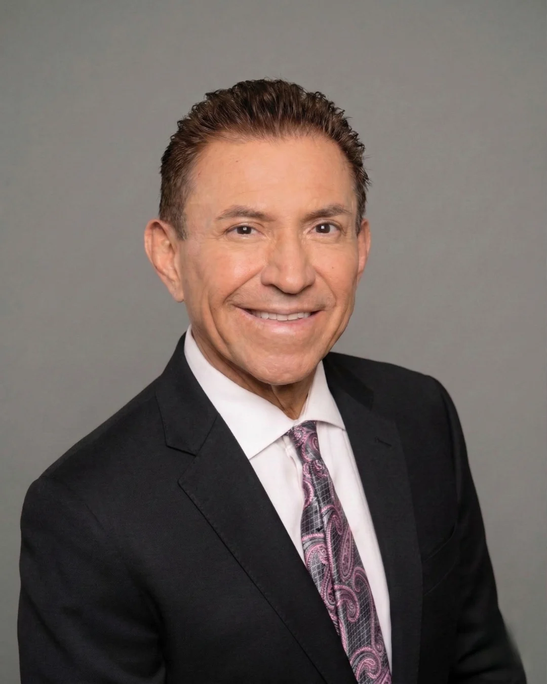 Smiling man in dark suit with paisley tie on plain gray background